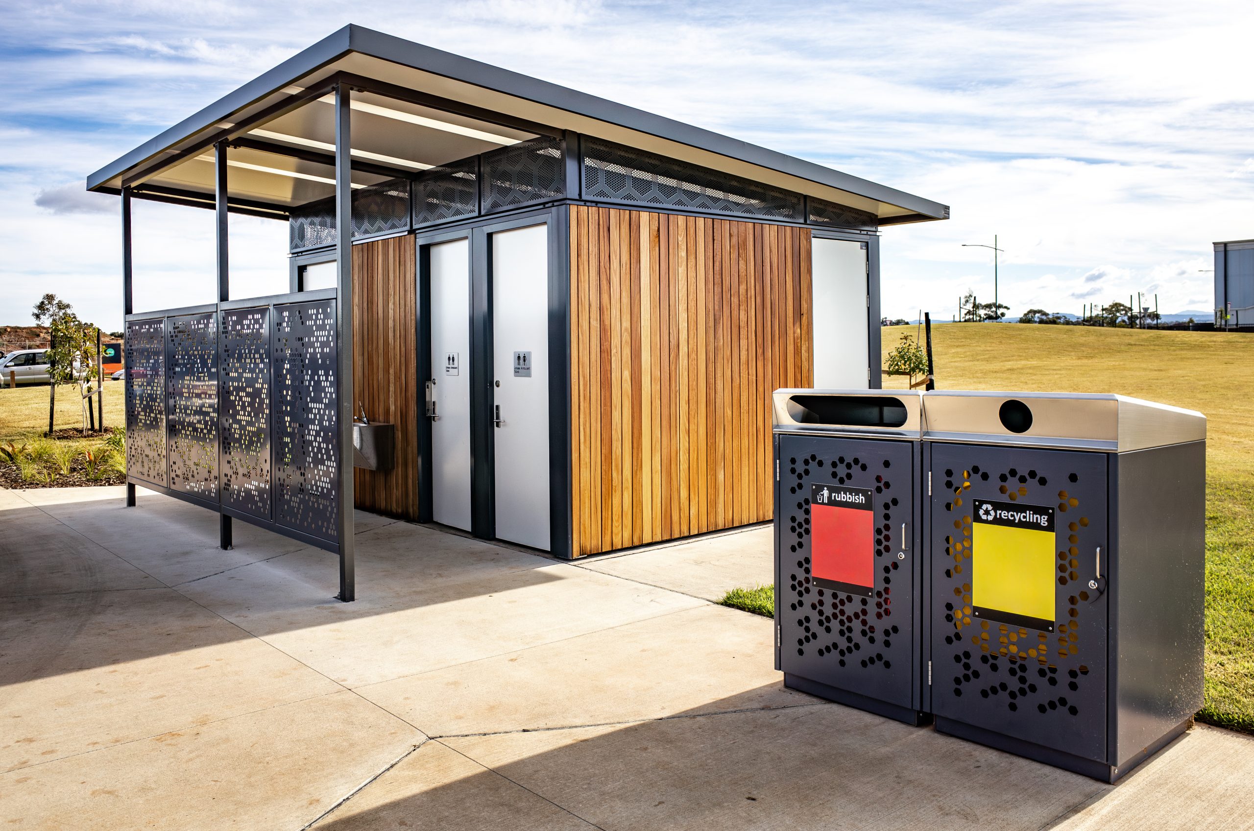 A public toilet block with wood paneling and perforated metal privacy screens, in a well-kept suburban park in Australia, accompanied by separate recycling and waste bins
