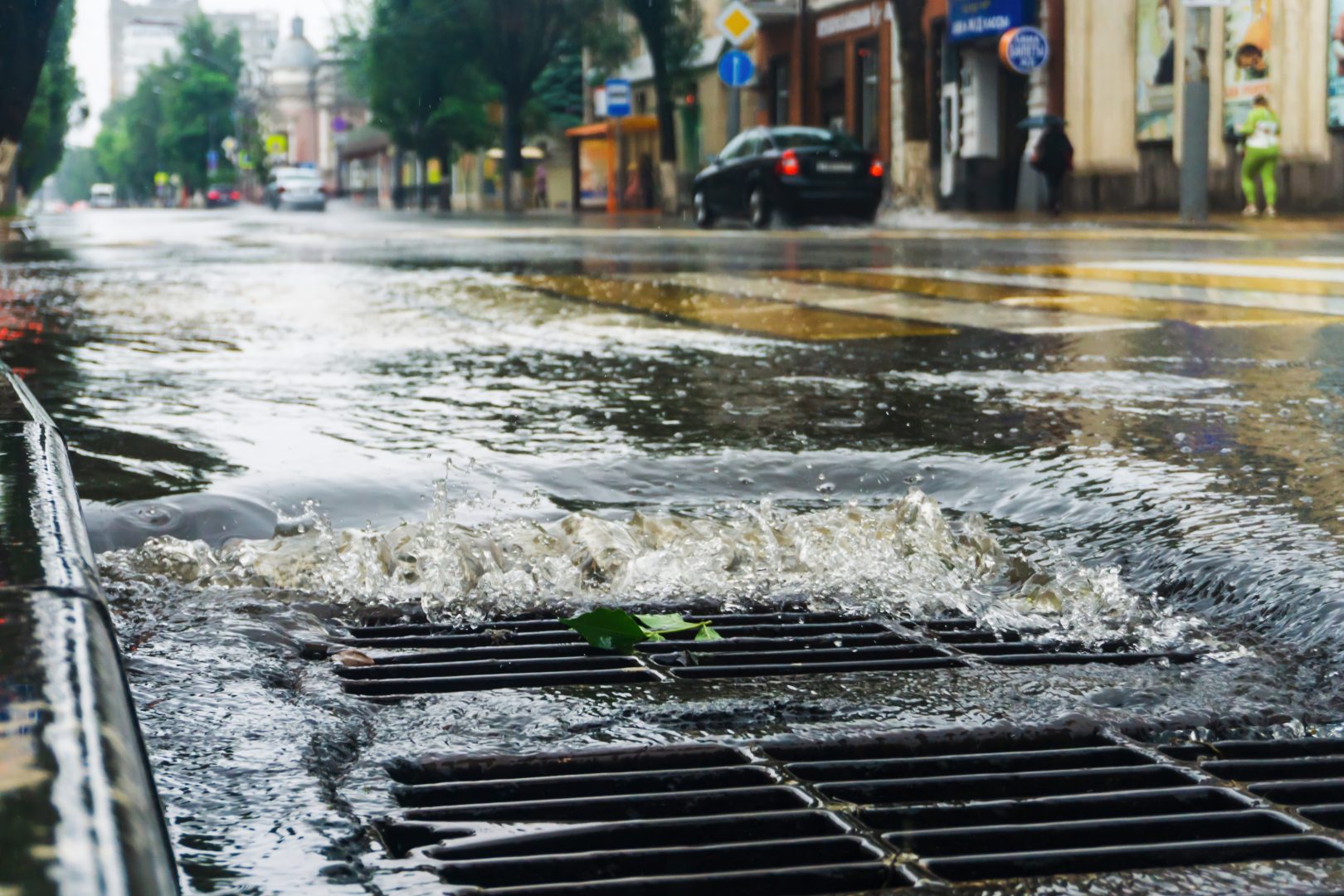 Close-up of stormwater pouring into a metal grate on a rainy urban street with traffic and buildings in soft focus.